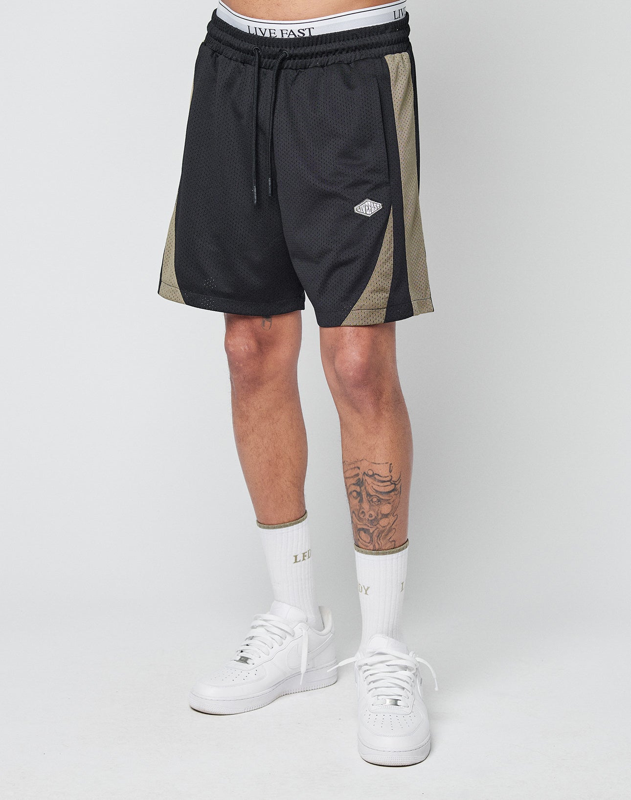 Wearing LIVE FAST DIE YOUNG Striped Mesh Shorts, white LIFE EASY crew socks, and white sneakers, a person with tattooed legs stands against a plain white background.