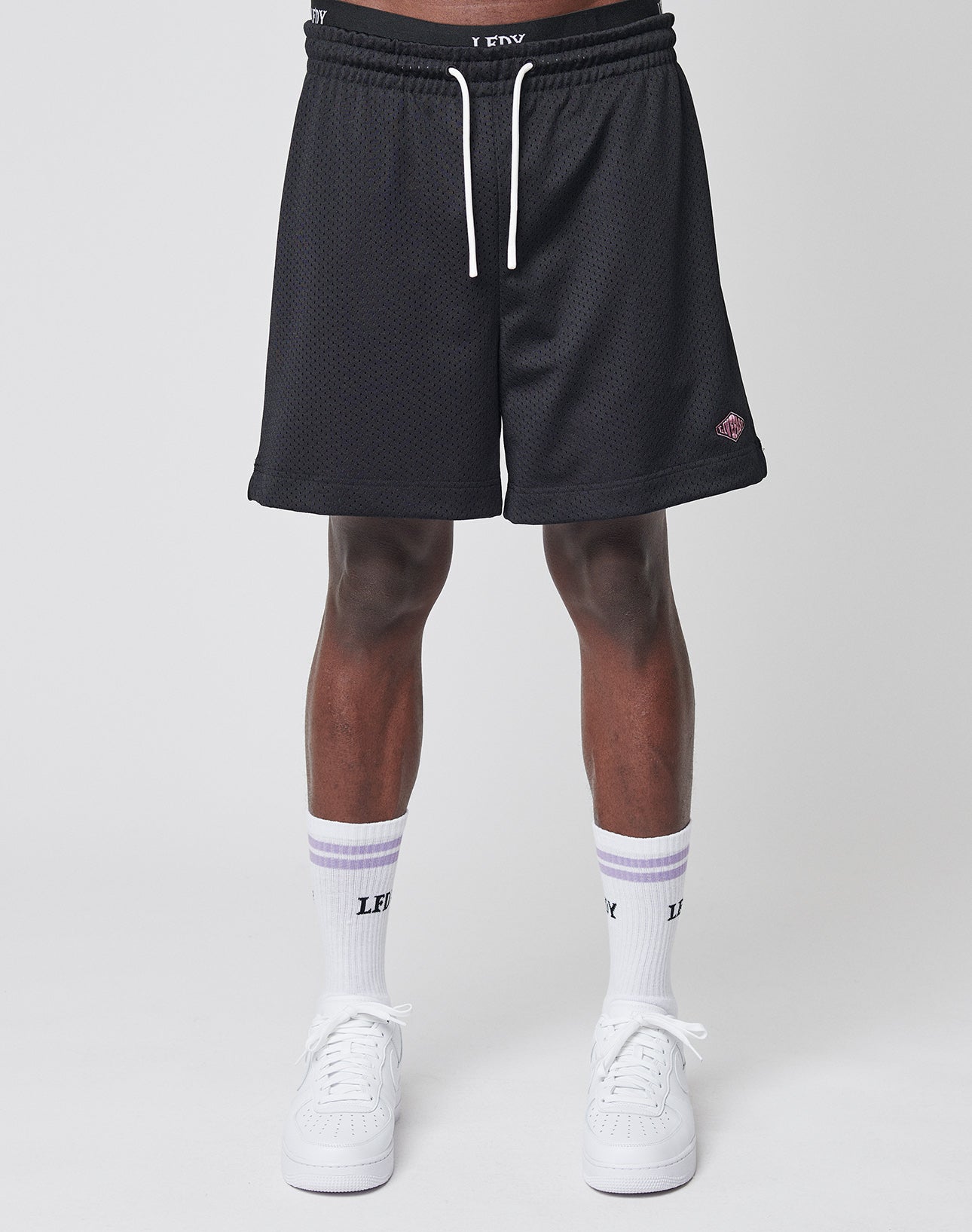 A person wears LIVE FAST DIE YOUNG Basic Mesh Shorts in black with an elastic waistband, white crew socks with LEYY text, and white sneakers against a plain light background. Image is cropped from the waist down.