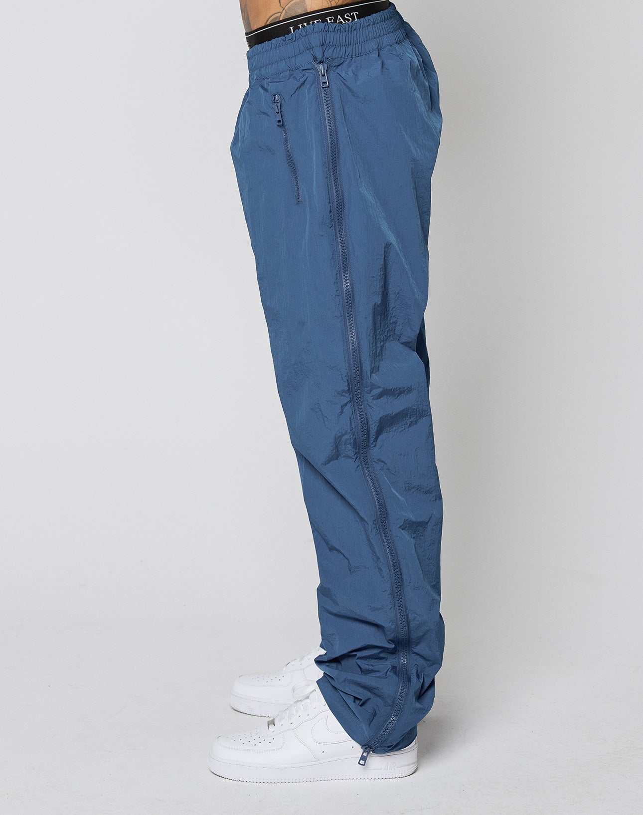 A person wears blue Zip Tape Track Pants by LIVE FAST DIE YOUNG with a black SAINT EASY waistband and white sneakers, standing against a plain white background.