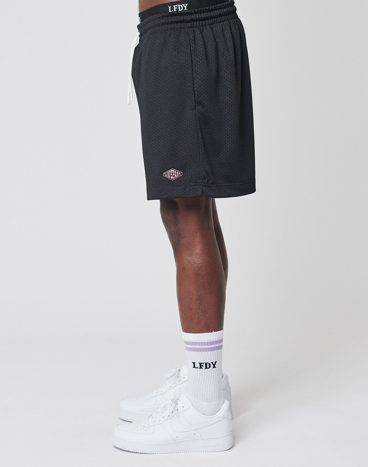 A person stands sideways, showing only their lower half, wearing LIVE FAST DIE YOUNG Basic Mesh Shorts in black with an elastic waistband, paired with white LFDY text socks and white sneakers against a plain white background.
