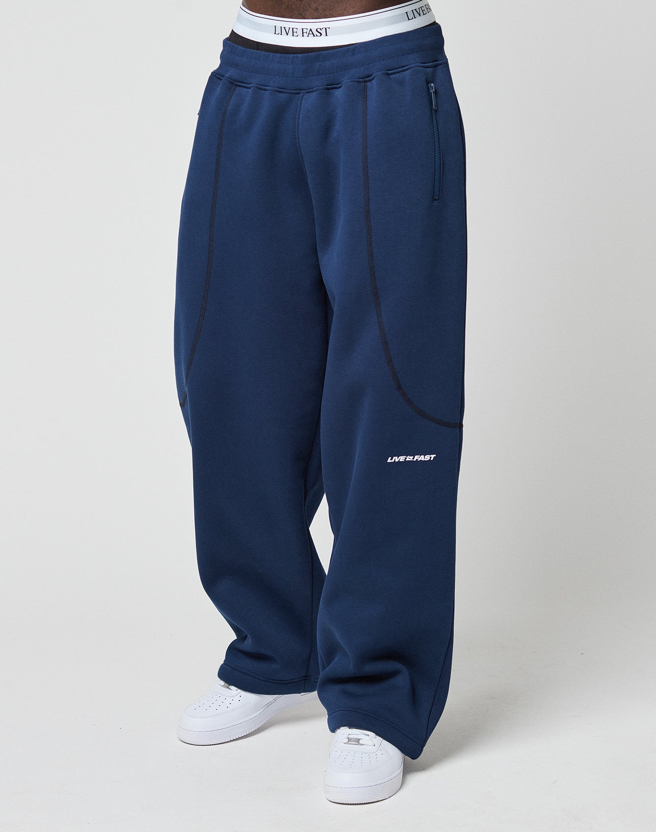 Someone wearing LIVE FAST DIE YOUNG’s LFDY Active Fleece Jogger in wide fit navy blue with black seam accents, logo print and LIVE FAST on the left thigh, paired with white sneakers and a visible LIVE FAST waistband.