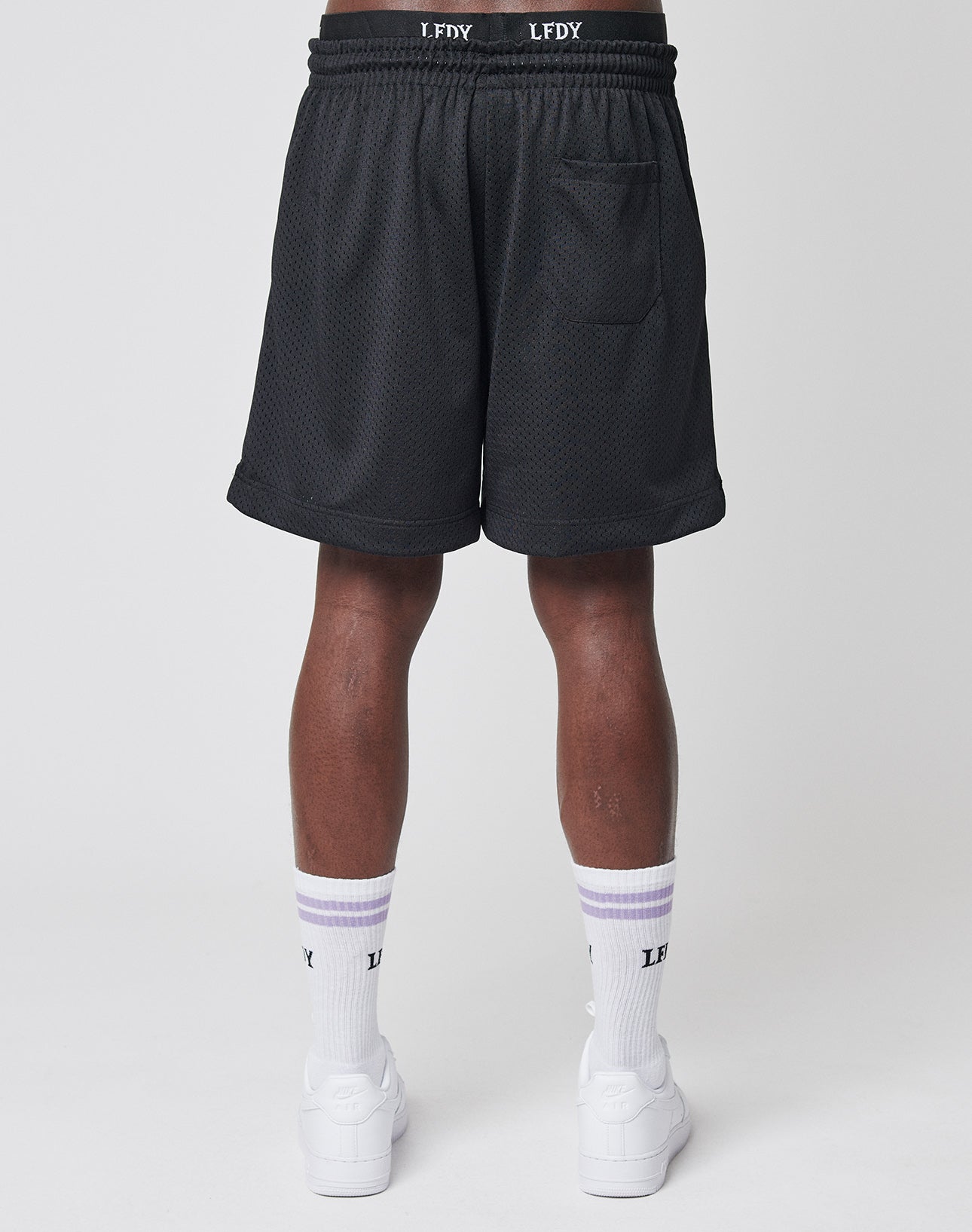 A person faces away, wearing LIVE FAST DIE YOUNG Basic Mesh Shorts with a back pocket and logo patch, white socks featuring purple stripes and LF DY text, and white sneakers against a plain background.
