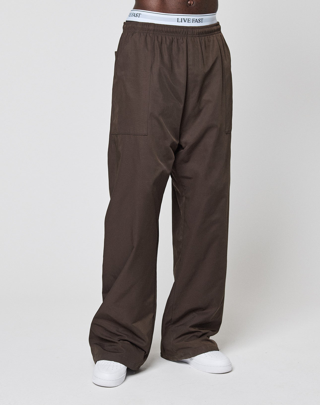 Model wears Baggy Nylon Pants in brown from LIVE FAST DIE YOUNG, featuring a wide fit, zip pockets, elastic waistband with exposed LIVE FAST branding, paired with white sneakers against a plain light background.
