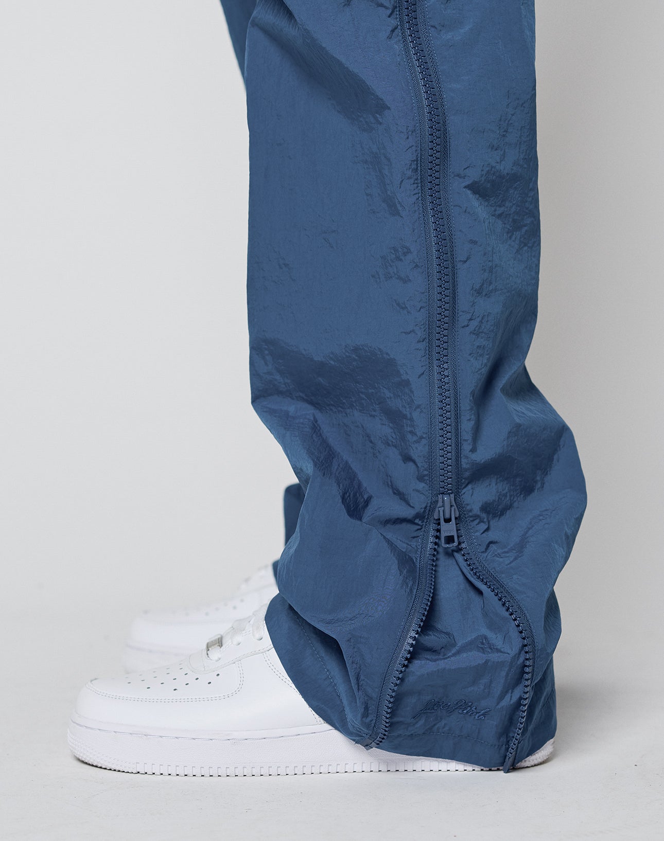 Close-up of a person wearing shiny blue LIVE FAST DIE YOUNG Zip Tape Track Pants with a two-way zipper along the side seam near the ankle, paired with white sneakers against a plain, light background.