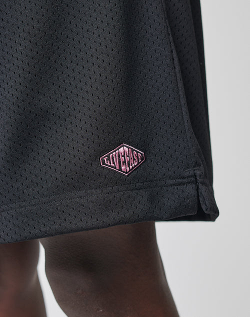 A close-up of LIVE FAST DIE YOUNG Basic Mesh Shorts in black polyester mesh with a pink diamond LIVECASH patch. These regular fit shorts have an elastischer Bund and show part of a persons leg below the hem.
