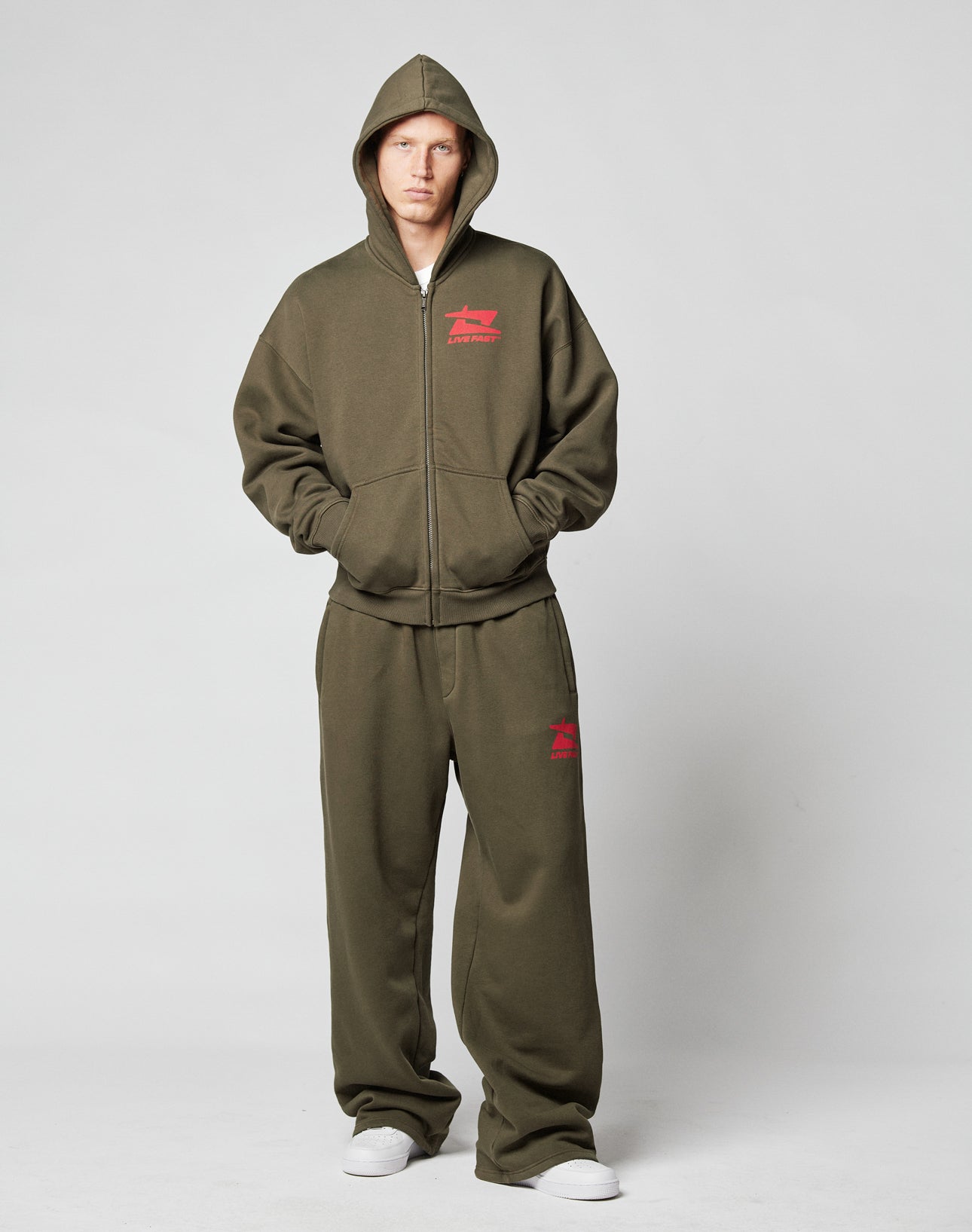 A person wears the LIVE FAST DIE YOUNG Live Fast Sweatsuit Jacket in olive green with a red front logo, hood, pockets, and white sneakersāan oversized fit made from premium cotton.