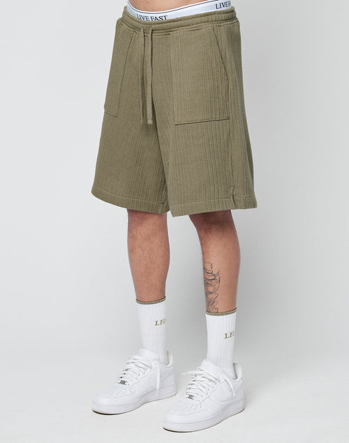 A person wears LIVE FAST DIE YOUNG Basic Knit Shorts in olive green with an elastic waistband, white LIVE FAST waistband underwear, white socks featuring LIVE FAST text, and white Nike sneakers against a plain light background.