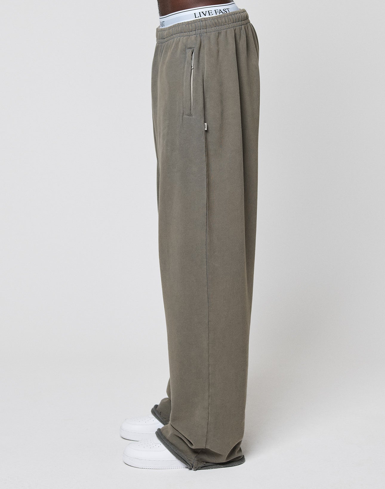 A person in LFDY Ripped Sweatpants by LIVE FAST DIE YOUNGāwide fit, olive green, 100% cotton, zipper pocket, white LIVE FAST waistbandāwith white sneakers, standing sideways against a plain white background.