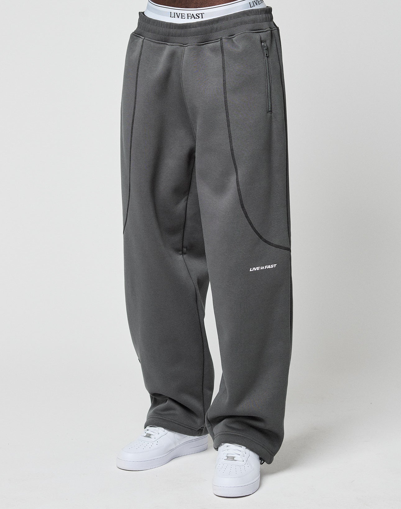 A person models the LIVE FAST DIE YOUNG LFDY Active Fleece Jogger in wide fit, dark gray, featuring zippered pockets and white LIVE FAST logos on the pocket area and waistband, styled with white sneakers against a plain background.