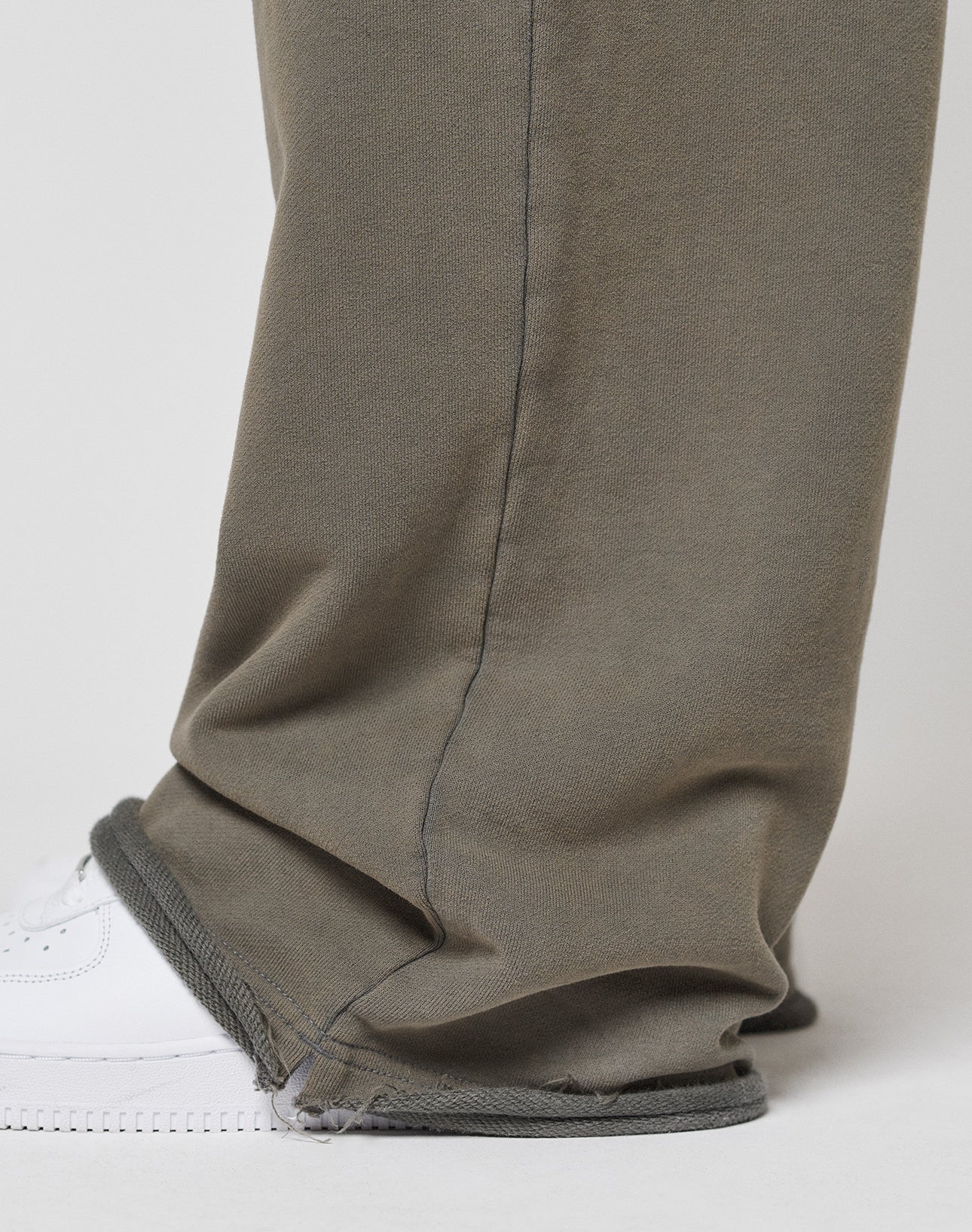 A close-up shows the lower leg in LIVE FAST DIE YOUNGās LFDY Ripped Sweatpantsāloose, olive-green with frayed hems and a Wide Fit, made from 100% Baumwolle with an Elastikbund, partially covering a white sneaker against a plain background.