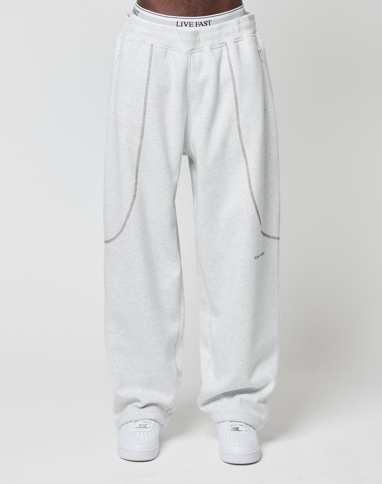 A person wears the LFDY Active Fleece Jogger by LIVE FAST DIE YOUNG—light gray, wide fit with seam details and a logo print waistband reading LIVE FAST—paired with white sneakers, against a plain white background.