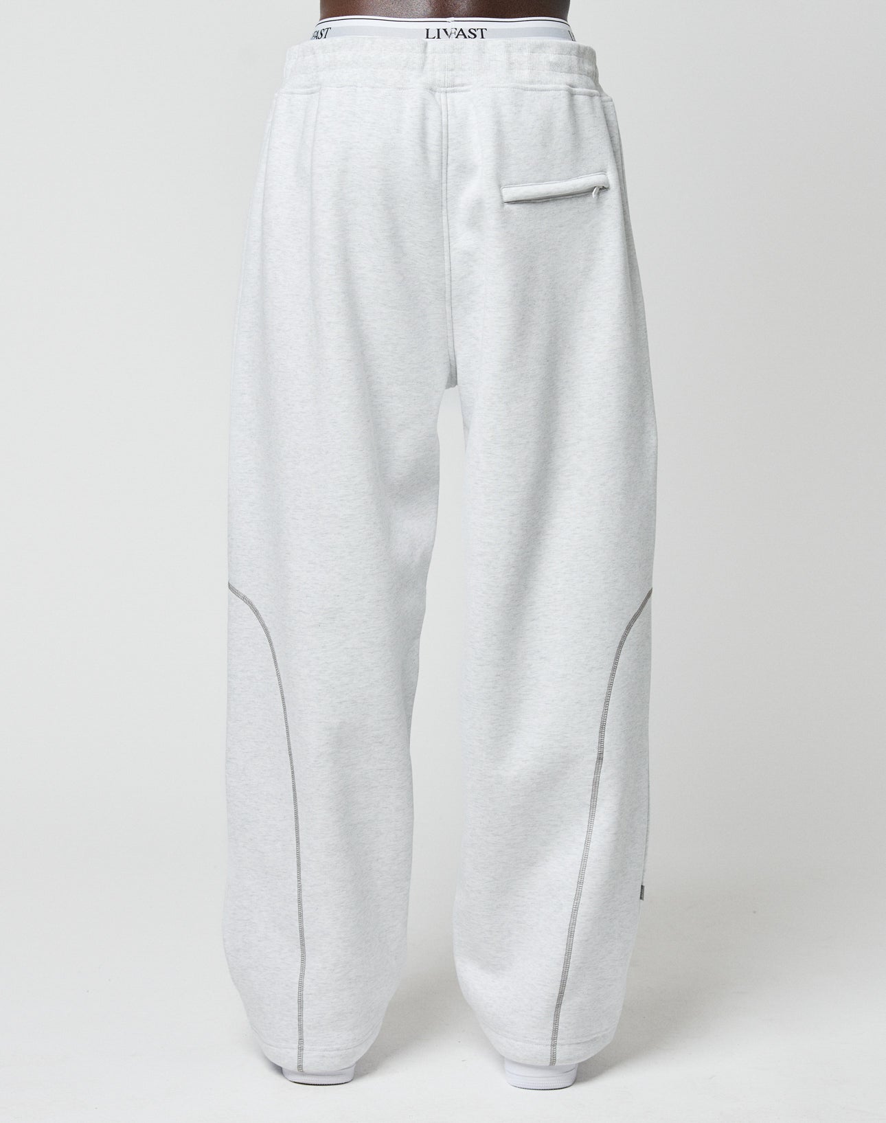Someone is seen from behind in LFDY Active Fleece Joggers by LIVE FAST DIE YOUNG. These light gray pants have a wide fit, right-side back pocket, curved seams, and a waistband with LINVAST logo print.