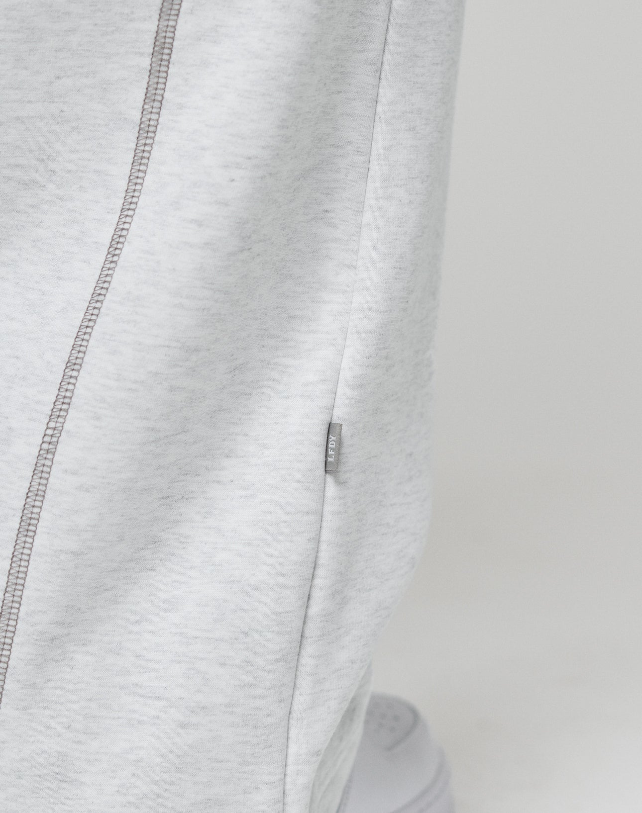 Close-up of LFDY Active Fleece Jogger by LIVE FAST DIE YOUNG in light gray, featuring a wide fit, visible side seam with contrast stitching, and a small branded fabric tag. The lower part of a white sneaker appears at the bottom.
