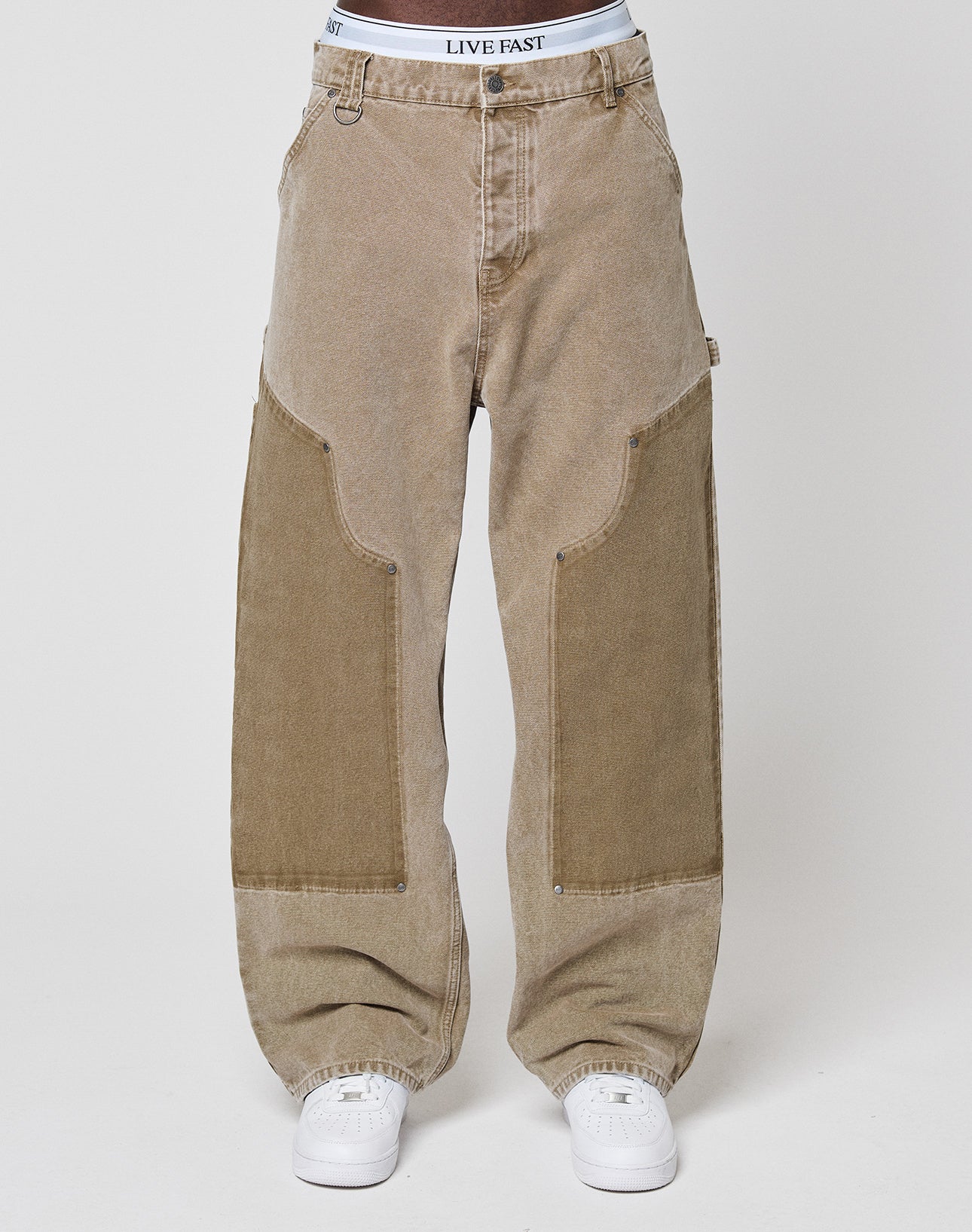 A person wears loose-fitting, light brown Faded Carpenter Pants by LIVE FAST DIE YOUNG with vintage wash, large dark brown thigh patches, white sneakers, and a visible LIVE FAST waistband against a plain, light backdrop.