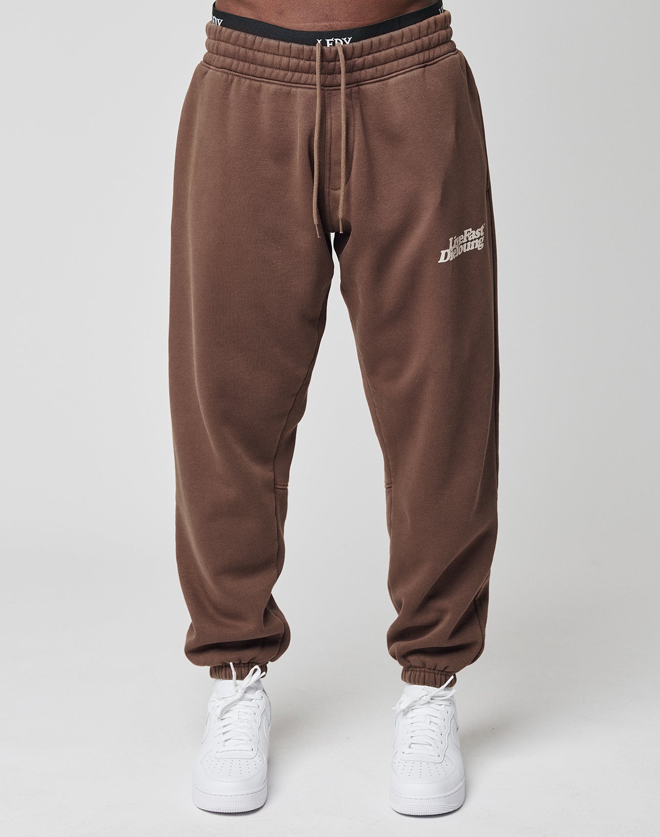 A person wears Basic Jogger sweatpants by LIVE FAST DIE YOUNG in brown with a regular fit, Daily Paper text on the upper left thigh, a wide elastic waistband, white sneakers, and visible black underwear against a plain light background.