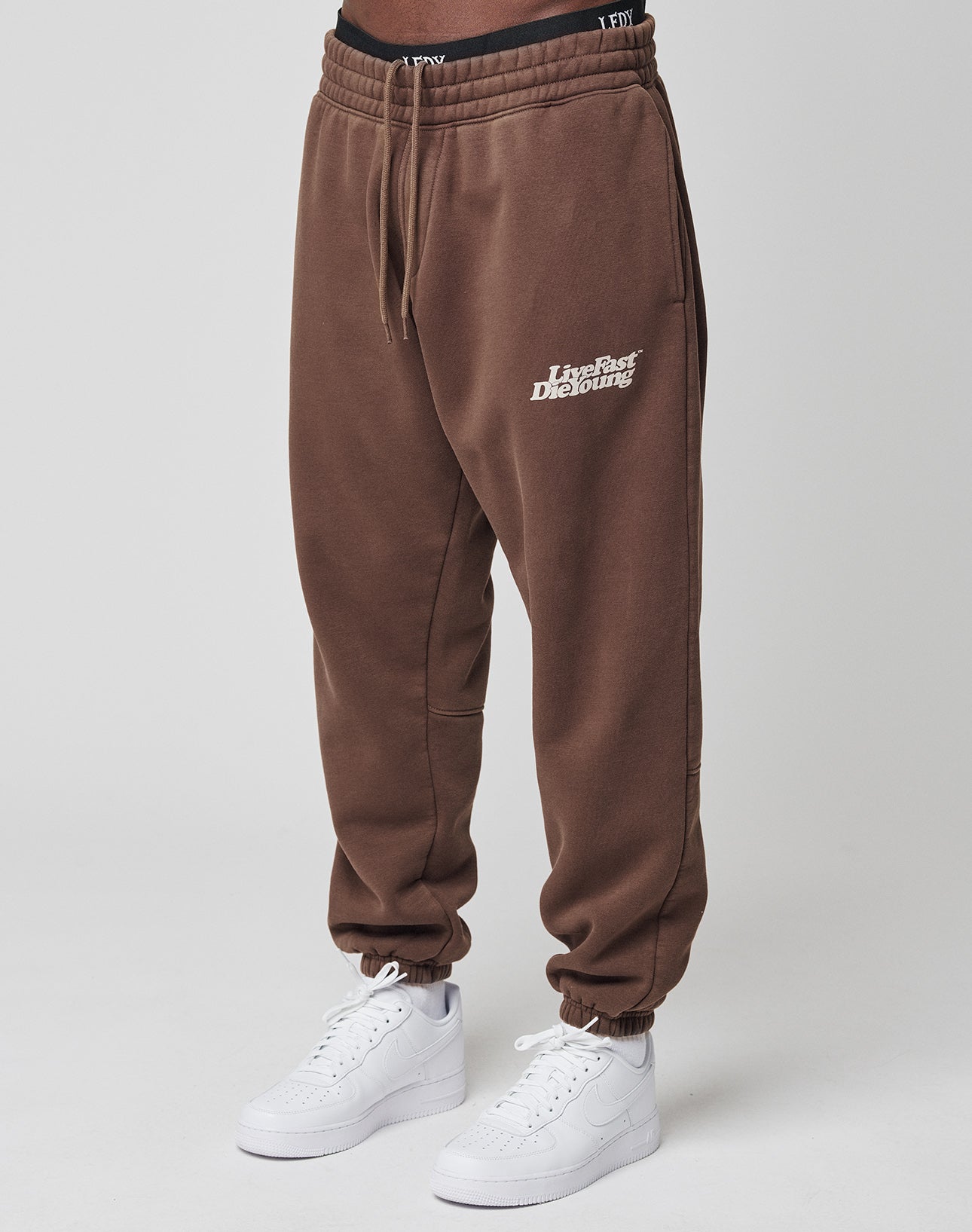 A person wears LIVE FAST DIE YOUNG’s Basic Jogger in brown with vintage wash and white embroidery on the left thigh, featuring a wide elastic waistband, styled with white sneakers and a visible black waistband against a neutral background.