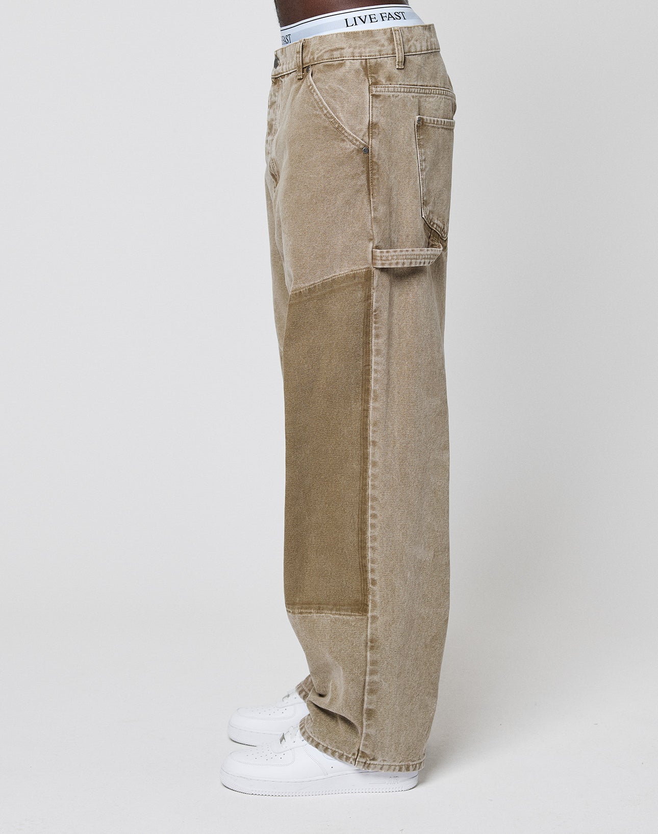 A person wearing LIVE FAST DIE YOUNG Faded Carpenter Pants with a light brown baggy fit, darker brown side patch, white sneakers, and a visible white LIVE FAST waistband stands against a plain white background.