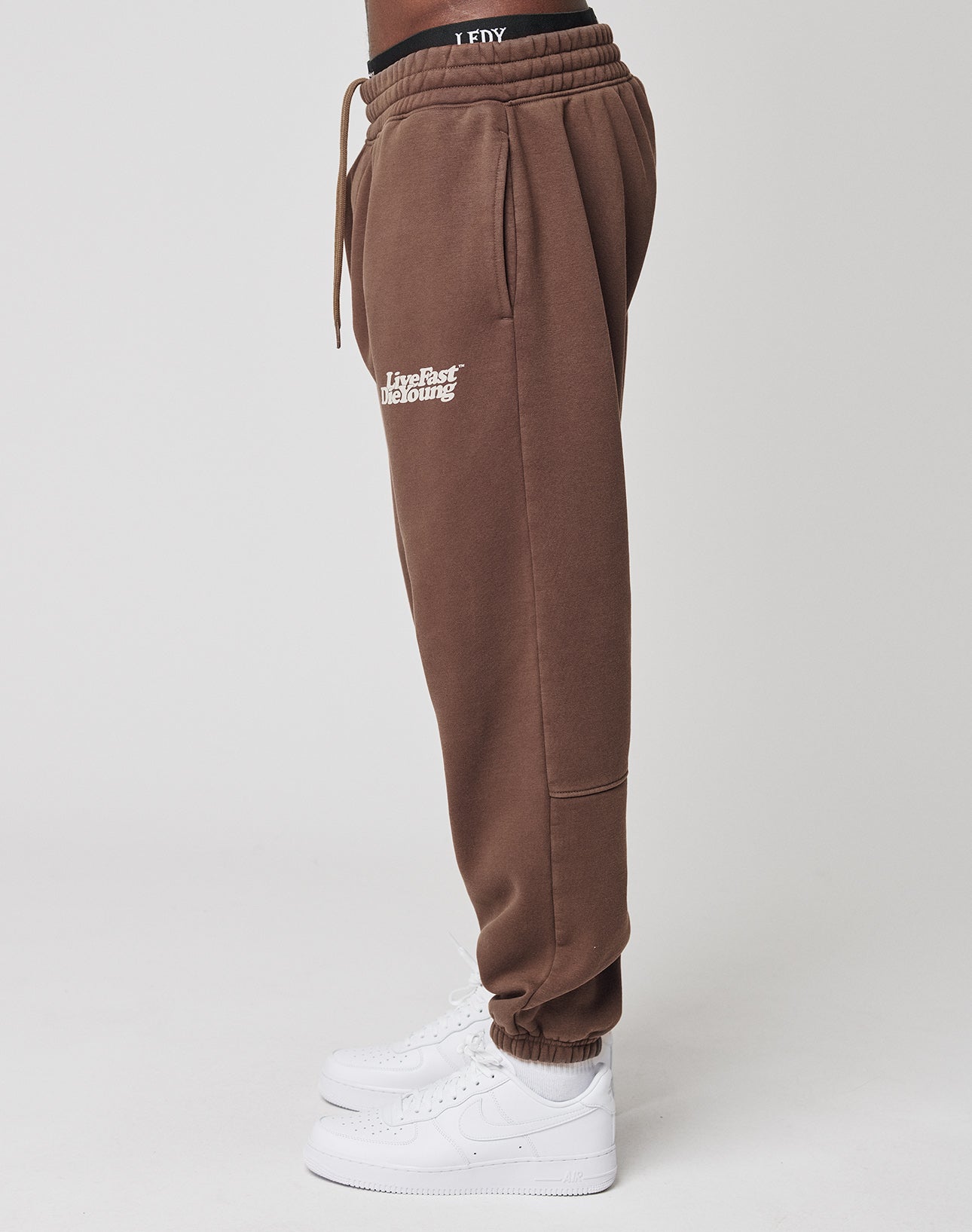 A person stands sideways in LIVE FAST DIE YOUNG Basic Jogger sweatpants—regular fit, brown, with a wider elastic waistband and white thigh text—paired with white sneakers and black I FLY waistband underwear. The background is plain white.