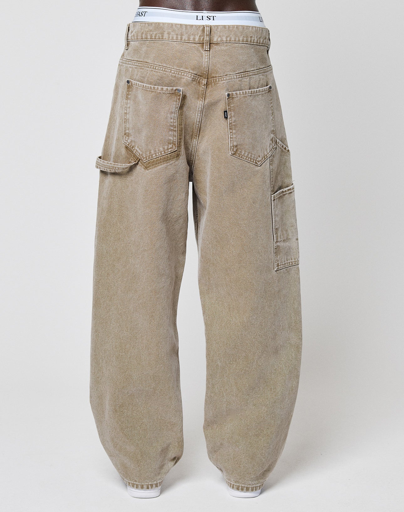 A person rocks the Carpenter Look in LIVE FAST DIE YOUNG Faded Carpenter Pants—baggy, light brown denim with large utility pockets. White LIST underwear peeks above the waist, adding a relaxed vintage vibe.