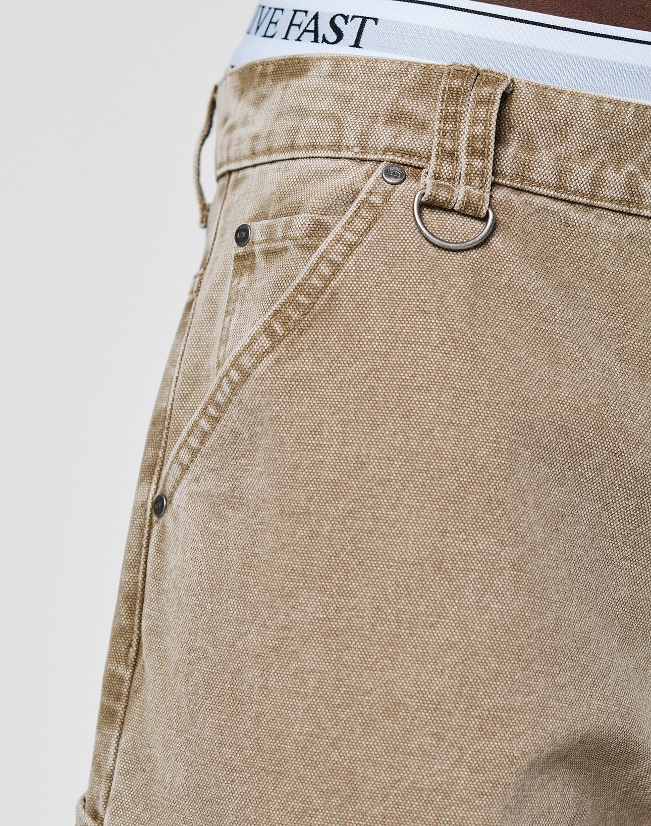 Close-up of LIVE FAST DIE YOUNG Faded Carpenter Pants in tan denim with vintage wash, metal rivets, and waistband loop ring. Baggy fit exposes white underwear band reading LIVE FAST. Set against a light plain background.