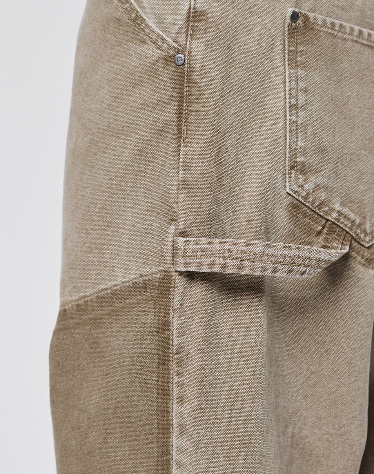 Close-up of Faded Carpenter Pants by LIVE FAST DIE YOUNG, featuring a baggy tan fit with diagonal front pocket, rivet, visible stitching, and thigh tool loop. The worn fabric adds to their rugged carpenter style.