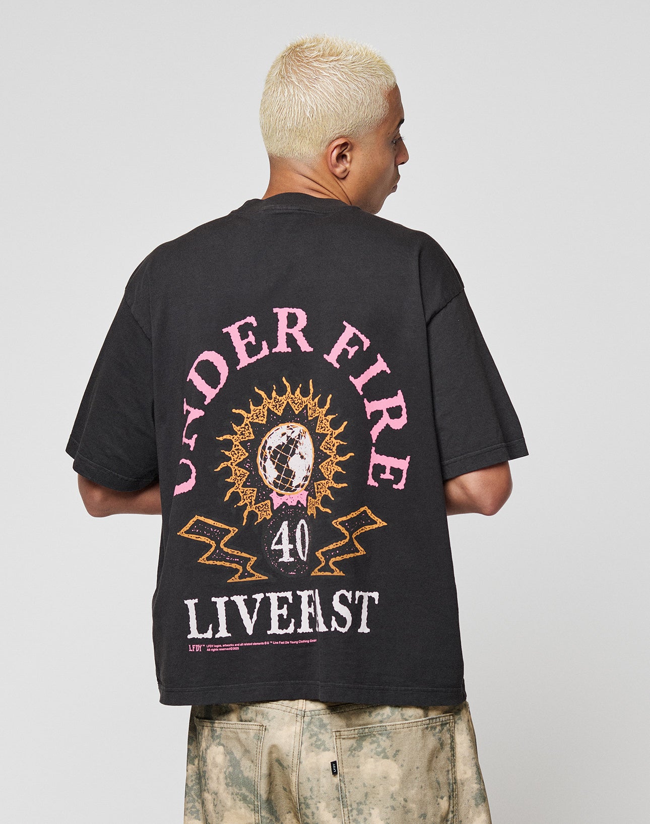 A person with short, bleached blonde hair wears the LFDY Under Fire Tee by LIVE FAST DIE YOUNG—an oversized black 100% cotton shirt with bold sunburst graphic, number 40, UNDER FIRE and LIVE FAST prints—standing with their back to the camera.
