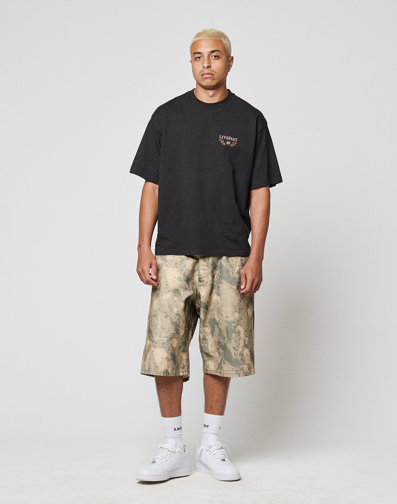 A person with short blonde hair wears the LIVE FAST DIE YOUNG LFDY Under Fire Tee, beige camo knee-length shorts, white socks, and white sneakers, standing against a plain white background.