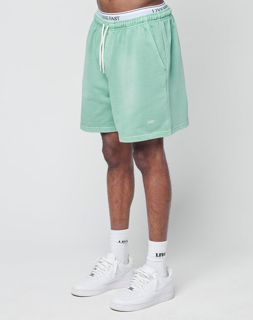 A person wears LIVE FAST DIE YOUNG Basic Sweat Shorts in mint green with handgefertigter Fade-Effekt, a white LIVE FAST waistband, LFDF white socks, and white sneakers while standing against a plain white background.