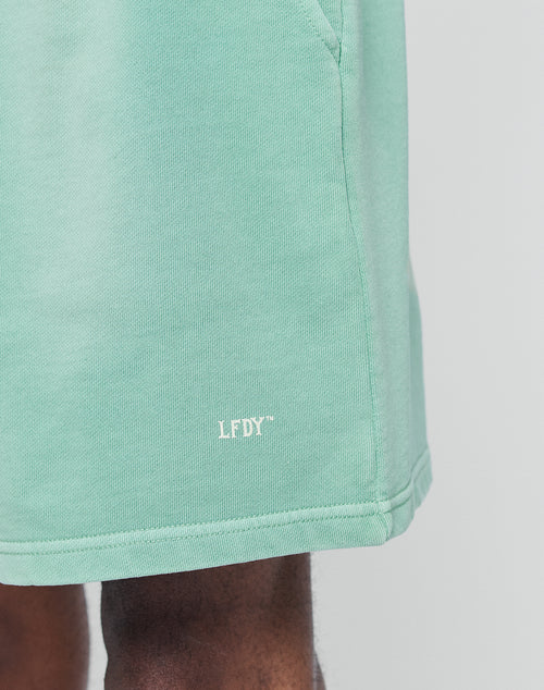 Close-up of a person wearing mint green Basic Sweat Shorts from LIVE FAST DIE YOUNG with white LFDY™ embroidery near the hem; their hand appears at the top left.