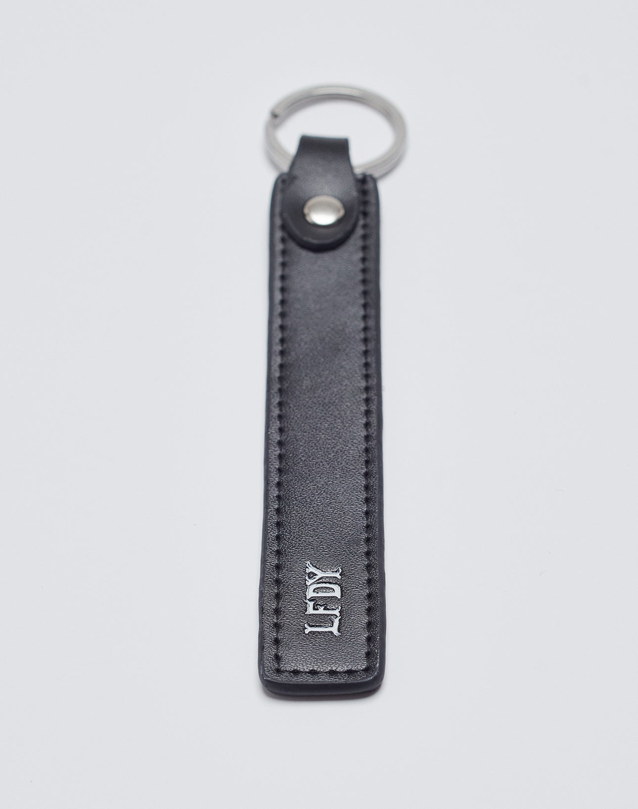 The Basic Keychain by LIVE FAST DIE YOUNG features black PU leather with visible stitching, a stainless steel ring, metal snap button, and white embossed “LDK” near the bottom for a stylish touch.