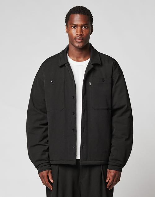 A man faces the camera in a LIVE FAST DIE YOUNG Padded Casual Shirt with chest pockets, styled oversized over a white tee and black pants, set against a plain light gray background.