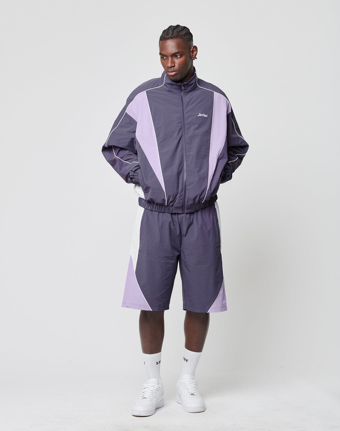 A man stands against a plain background, wearing LIVE FAST DIE YOUNG Basic Nylon Trackshorts and a matching Regular Fit purple and lavender tracksuit jacket with elastic cuffs and logo embroidery. His hands are in his pockets as he looks slightly downward.