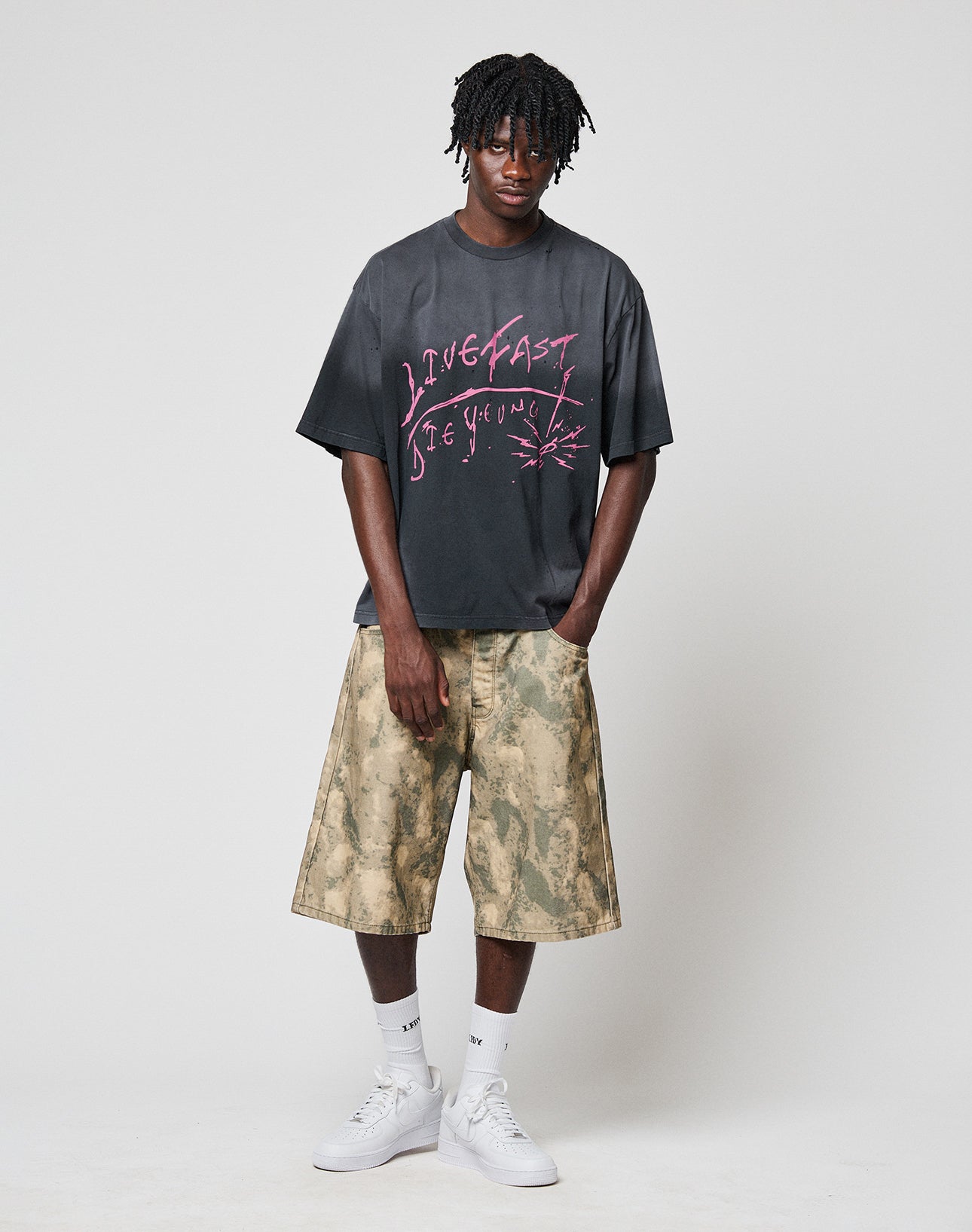 A man stands against a plain background wearing the LFDY Scribble Tee by LIVE FAST DIE YOUNG, tan tie-dye knee-length shorts, white crew socks, and white sneakers. His hands are in his shorts’ pockets.