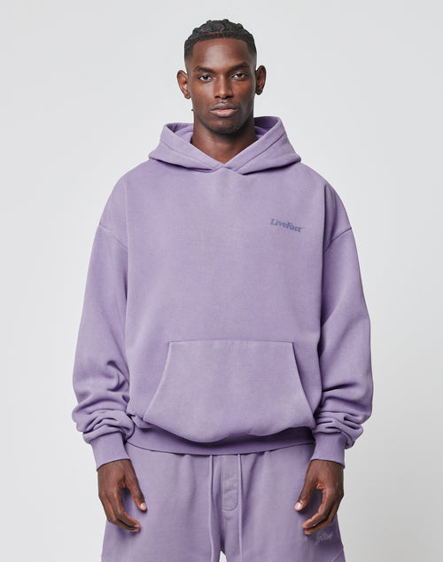 A man faces forward in an oversize, light purple Basic 360 Hooded and matching sweatpants by LIVE FAST DIE YOUNG, both featuring LiveFast text, made from Baumwolle Polyester, against a plain white background.