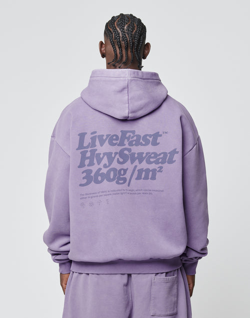 Seen from behind, a person with braided hair wears the LIVE FAST DIE YOUNG Basic 360 Hooded in Oversize Fit, lavender, with bold back text LiveFast HvySweat 360g/m² and smaller print below. Vintage-washed Baumwolle-Polyester blend.