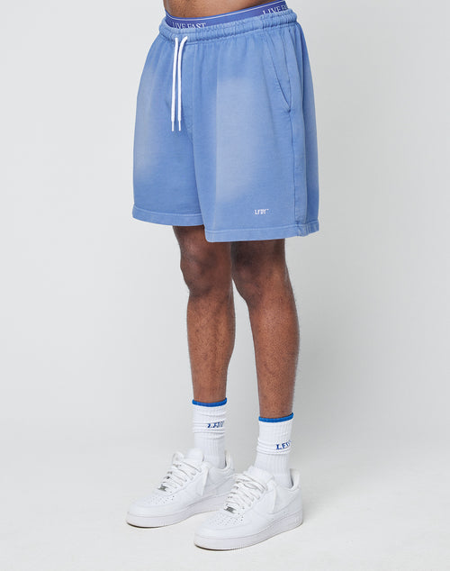A person wearing LIVE FAST DIE YOUNG Basic Sweat Shorts in light blue with white drawstrings, an elastic waistband, and a small LIFT logo near the hem stands against a plain light background, paired with white socks with blue detail and white sneakers.