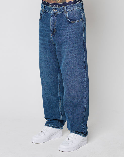 Model wears Wide Denim by LIVE FAST DIE YOUNG—loose-fitting blue jeans made from 100% cotton—with white sneakers and a branded waistband reading LIVE FAST, standing against a plain white background.