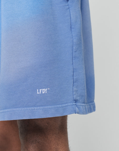 Close-up of a person wearing LIVE FAST DIE YOUNG’s Basic Sweat Shorts in blue, featuring white embroidered LFDY text near the hem and a regular fit, set against a plain light background.