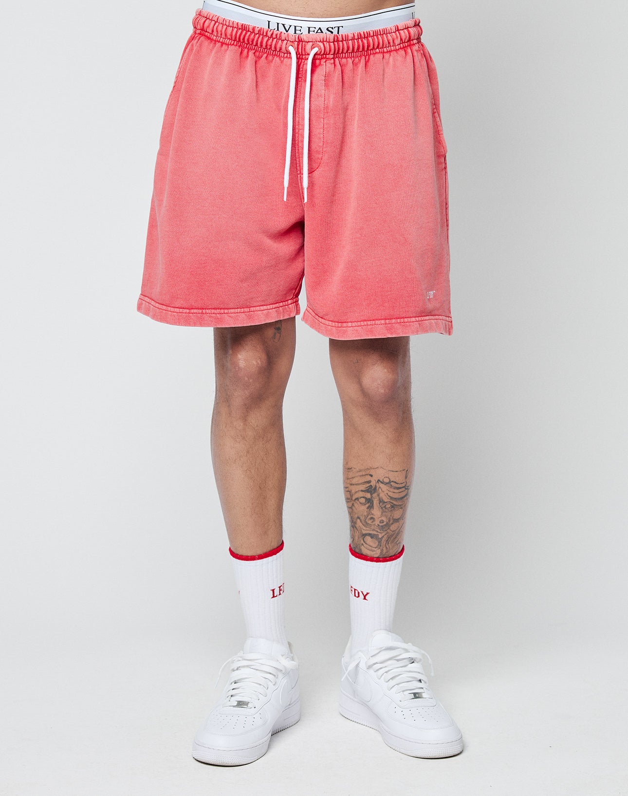 A person wears LIVE FAST DIE YOUNG Basic Sweat Shorts in pink with a handmade fade effect, white crew socks with red details, and white sneakers. Only their legs and part of their torso are visible against a plain light background.