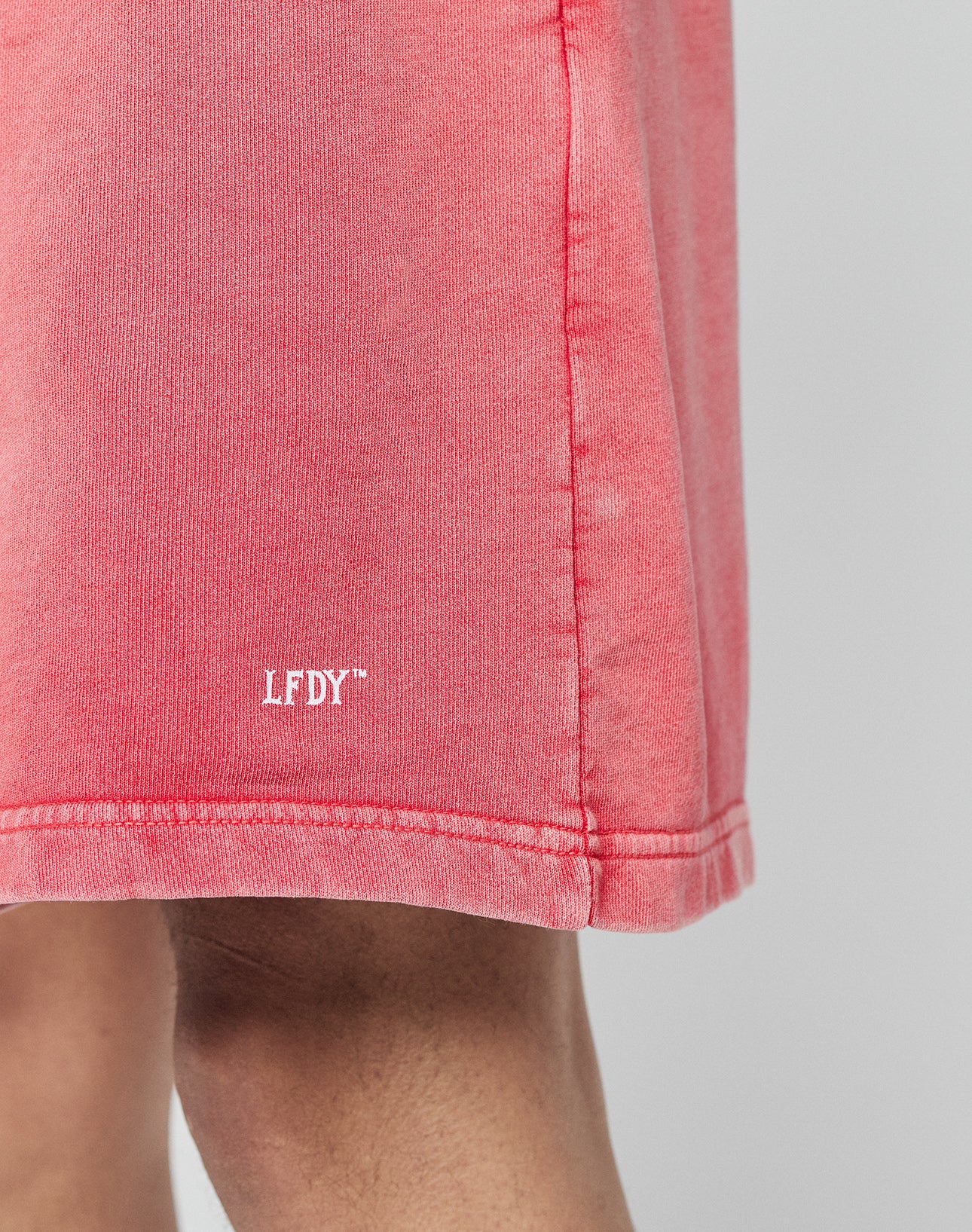 A close-up of a person wearing LIVE FAST DIE YOUNG Basic Sweat Shorts in pink, featuring an elastic waistband and a white LFDY™ logo near the hem. Only part of the leg and the subtly faded lower shorts are visible.