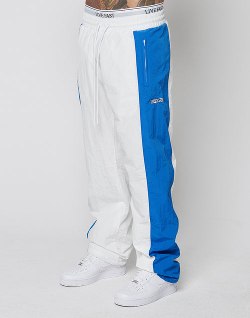 A person wears white LIVE FAST DIE YOUNG LFDY Trackpants 3000 with bright blue side panels and LIVE FAST elastic waistband, paired with white sneakers and showing tattoos, standing against a white background.
