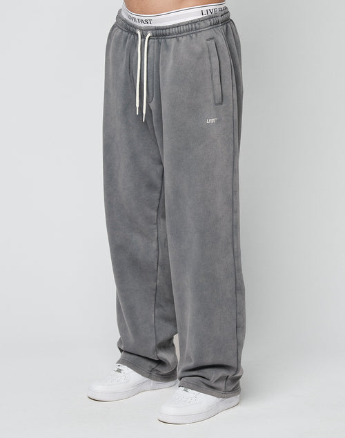 A person wears Basic Wide Jogger sweatpants by LIVE FAST DIE YOUNG in gray, with an elastic waistband with drawstring and LIFE print on the left thigh, plus a LIVE FAST logo on the waistband, paired with white sneakers against a plain background.