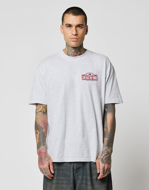 A tattooed man wears the LIVE FAST DIE YOUNG Live Fast 2013 Tee, a regular fit light gray T-shirt made from 100% Baumwolle, featuring a small red and white LIVES FAST DIE YOUNG graphic on the chest, against a plain background.