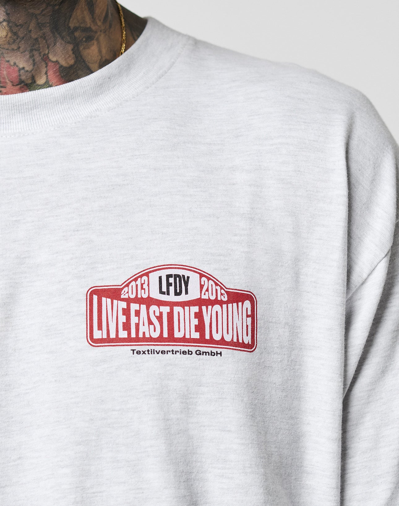 A close-up of the light gray Live Fast 2013 Tee by LIVE FAST DIE YOUNG, featuring a red and white chest logo. A tattooed neck and gold necklace are partially visible. Made from 100% Baumwolle (cotton).