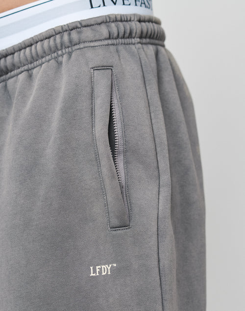Close-up of a person wearing the Basic Wide Jogger by LIVE FAST DIE YOUNG, showing an elastic waistband with drawcord, zippered pocket with logo print, and white underwear waistband with LIVE FAST text peeking above the pants.
