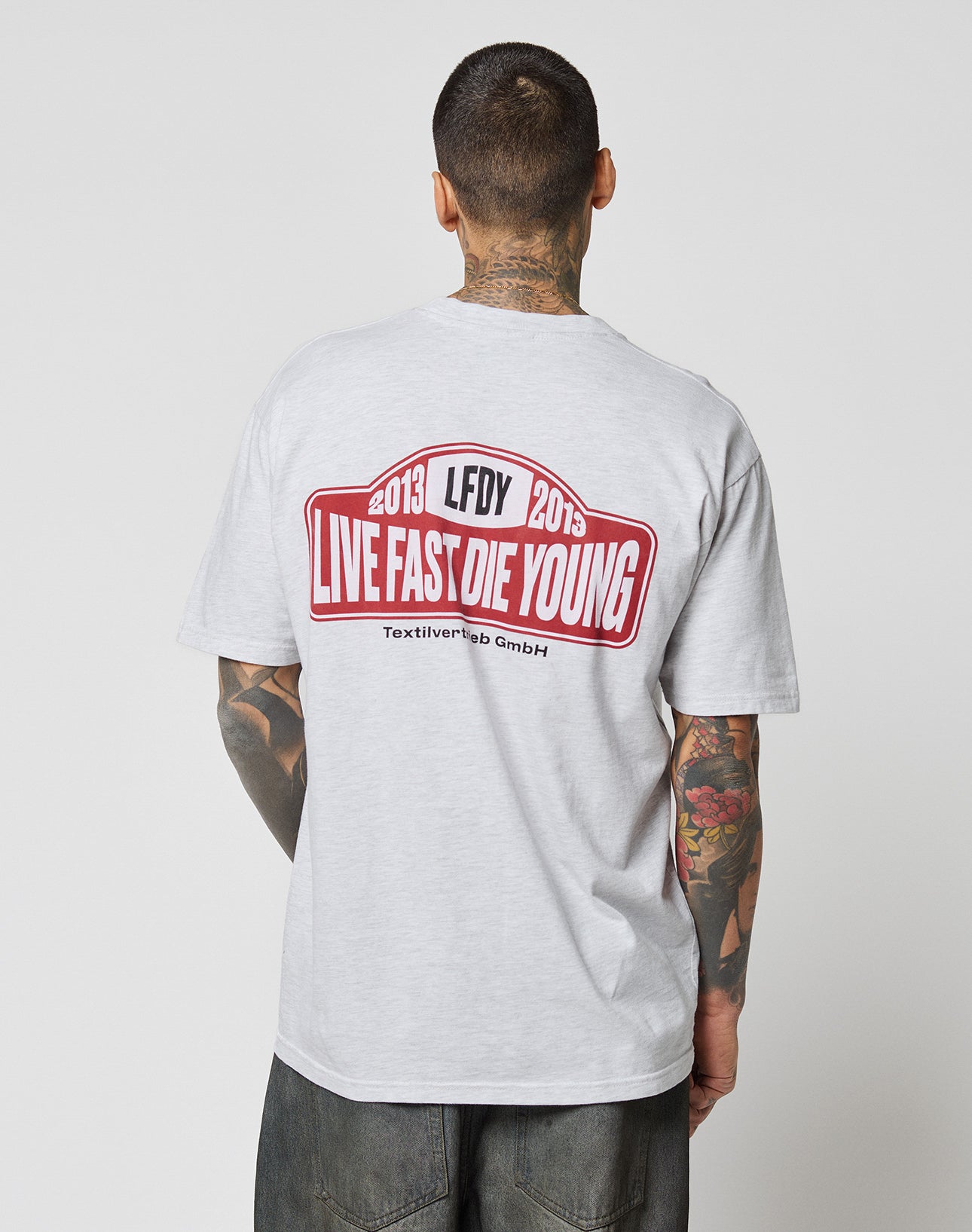 A tattooed person, seen from behind, wears the LIVE FAST DIE YOUNG Live Fast 2013 Tee—a light gray Regular Fit T-shirt made of 100% cotton with a bold red and white graphic and the dates 2013 and 2015 on the back.