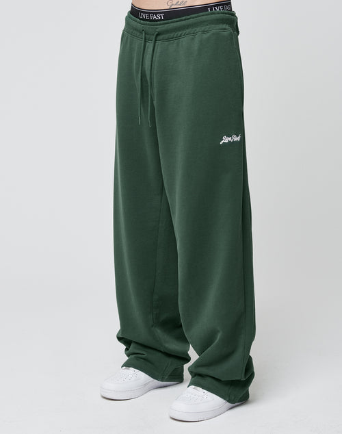 A person wears LIVE FAST DIE YOUNG’s Basic Wide Jogger in dark green with Live Well embroidery, an elastic waistband, and white sneakers. The waistband of their black LIVE FAST DIE YOUNG underwear is visible above the pants.