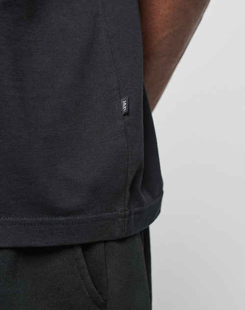 Close-up of a person wearing the LIVE FAST DIE YOUNG Basic Heavy Tee in black, styled with black pants. A small black tag with white text is sewn into the tee’s side seam. Only the lower torso and part of the arm are visible.
