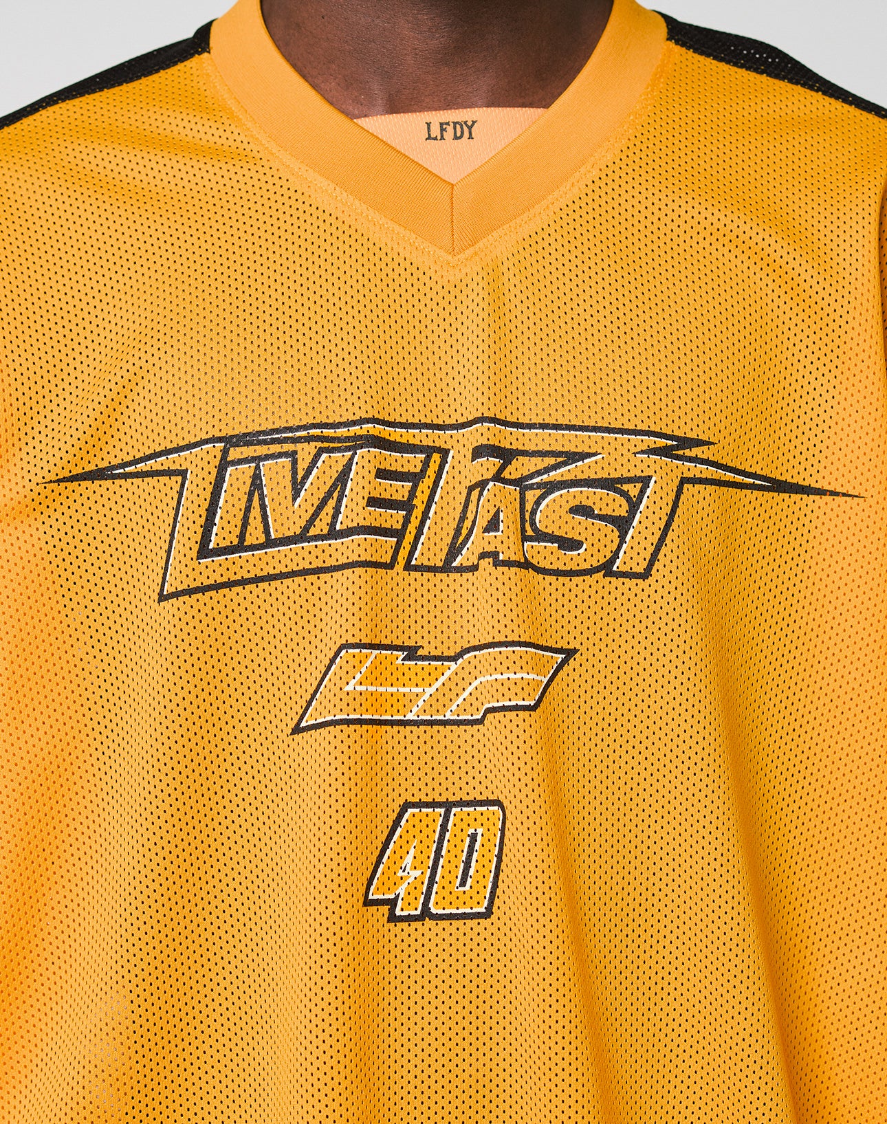 A close-up of a person wearing the LFDY Autobahn Jersey by LIVE FAST DIE YOUNG, featuring a yellow mesh polyester fabric, oversize fit, black “LIVE FAST” and “GF 40” prints on the front, and multilogo details including LFDY on the neckline.