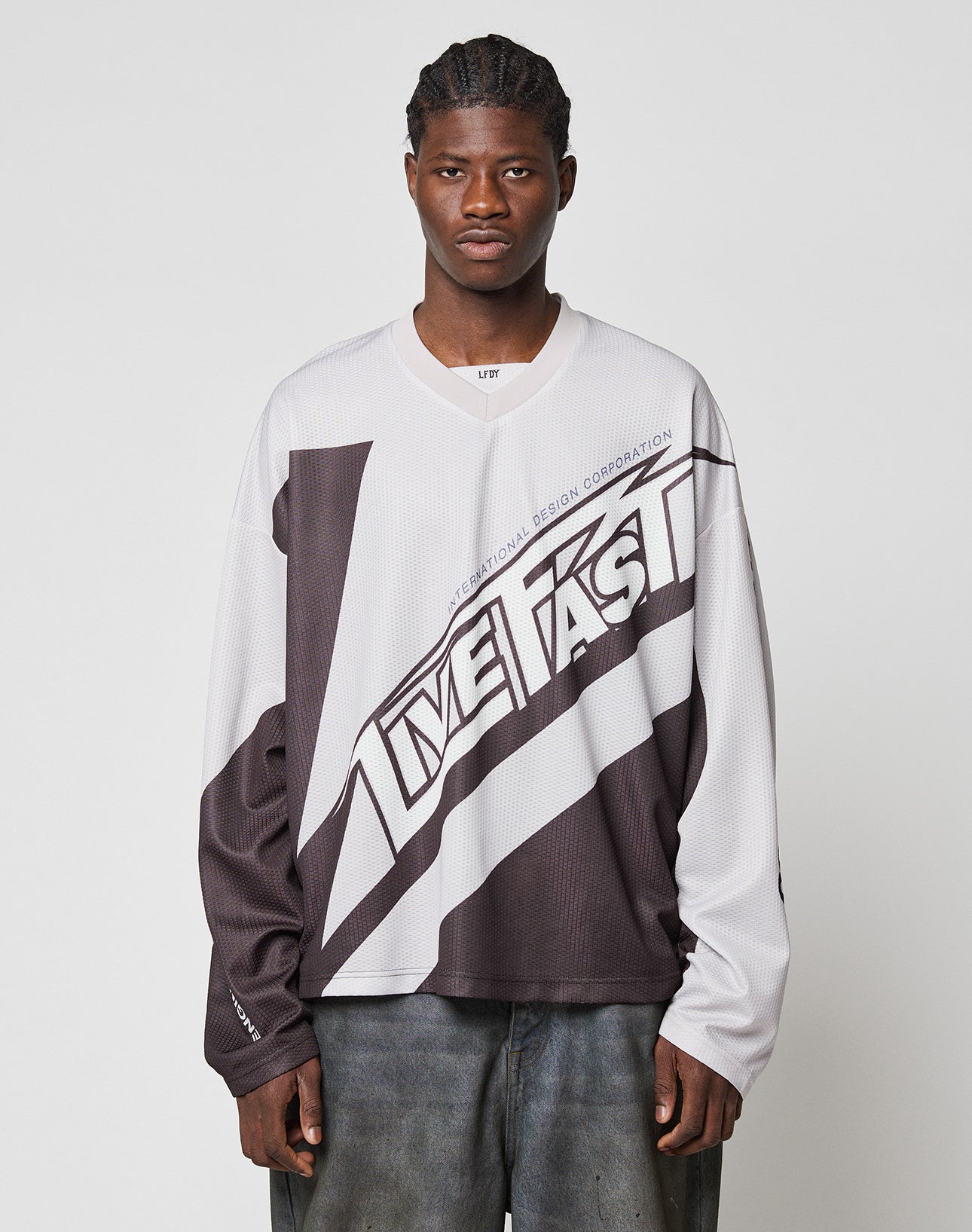 A man with braided hair wears an oversize, long-sleeve black and white LFDY Racetrack Jersey by LIVE FAST DIE YOUNG, standing against a plain light gray background.